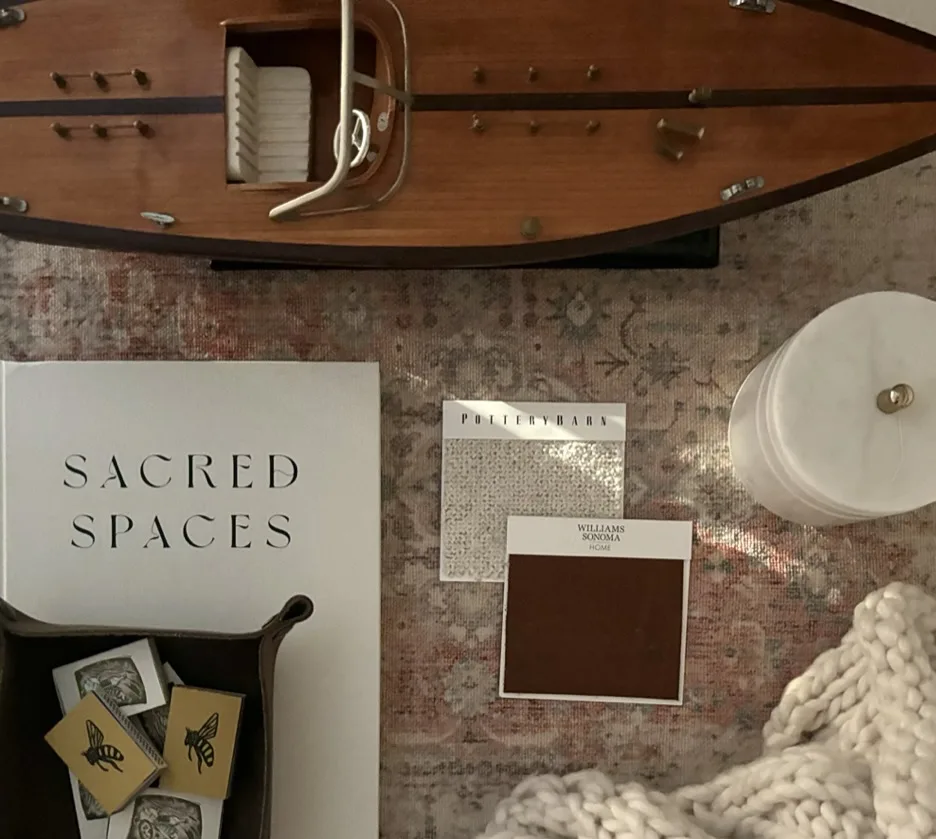 Styled vignette with model sailboat, "Sacred Spaces" design book, and fabric swatches on a vintage rug