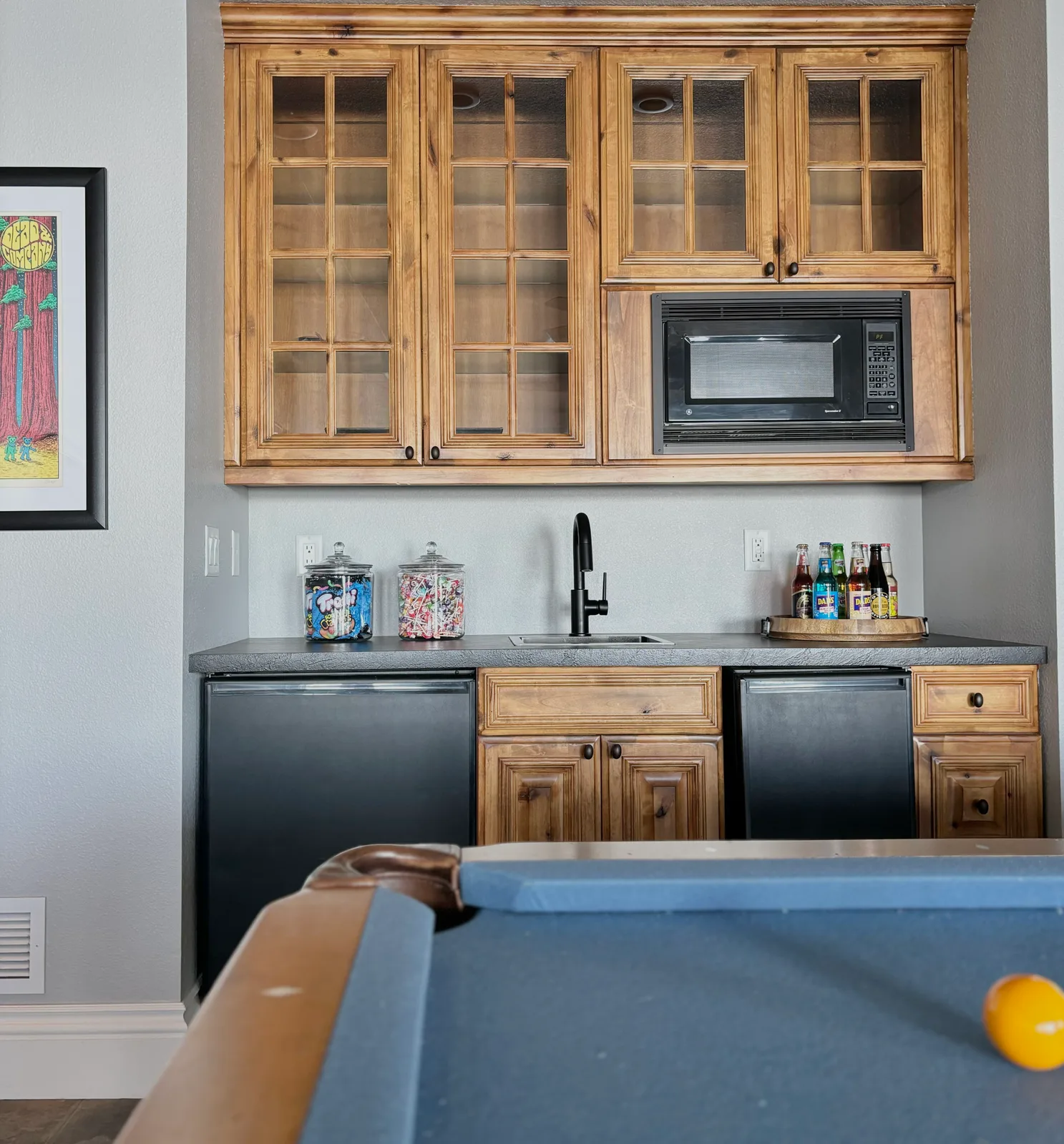 Rustic pine glass-front bar cabinet with candy jars and black mini fridges beside the game room