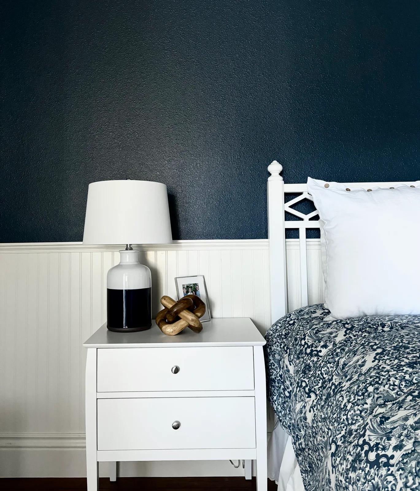 White bedside table with chrome lamp and wooden bear figurine against navy beadboard wall