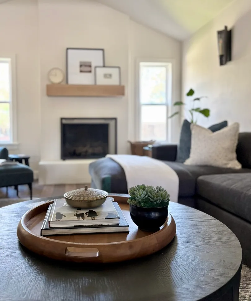 Round wood tray with framed art and fireplace in background, dark sectional and vaulted ceiling