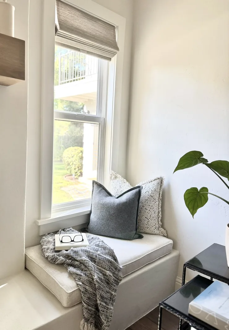 Window seat reading nook with built-in cushion, throw blanket, woven shade, and trailing plant