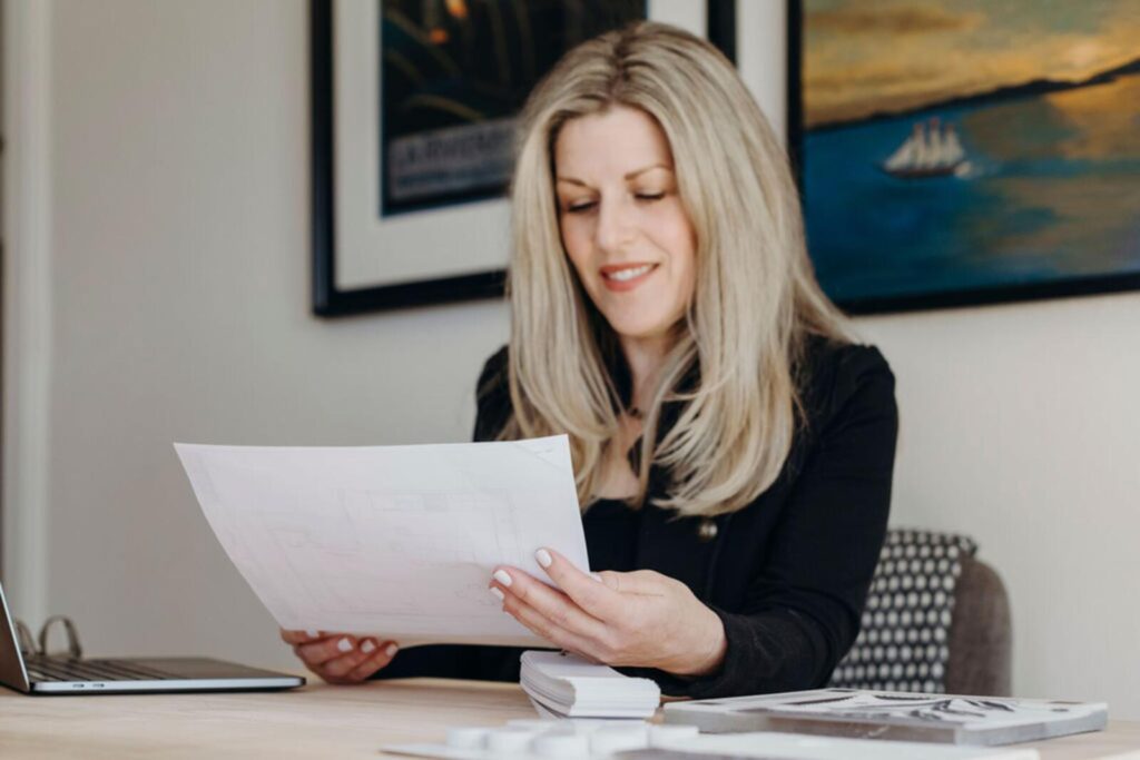 Modern workspace featuring a woman reviewing architectural plans on a wooden desk. The room has framed artworks on the walls