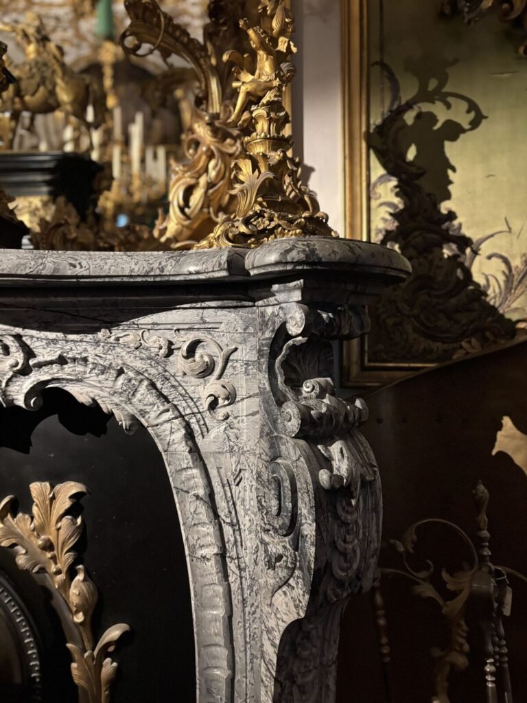 Ornate baroque-style fireplace mantel with intricate carvings and a mix of marble and gilded accents, set against a richly de
