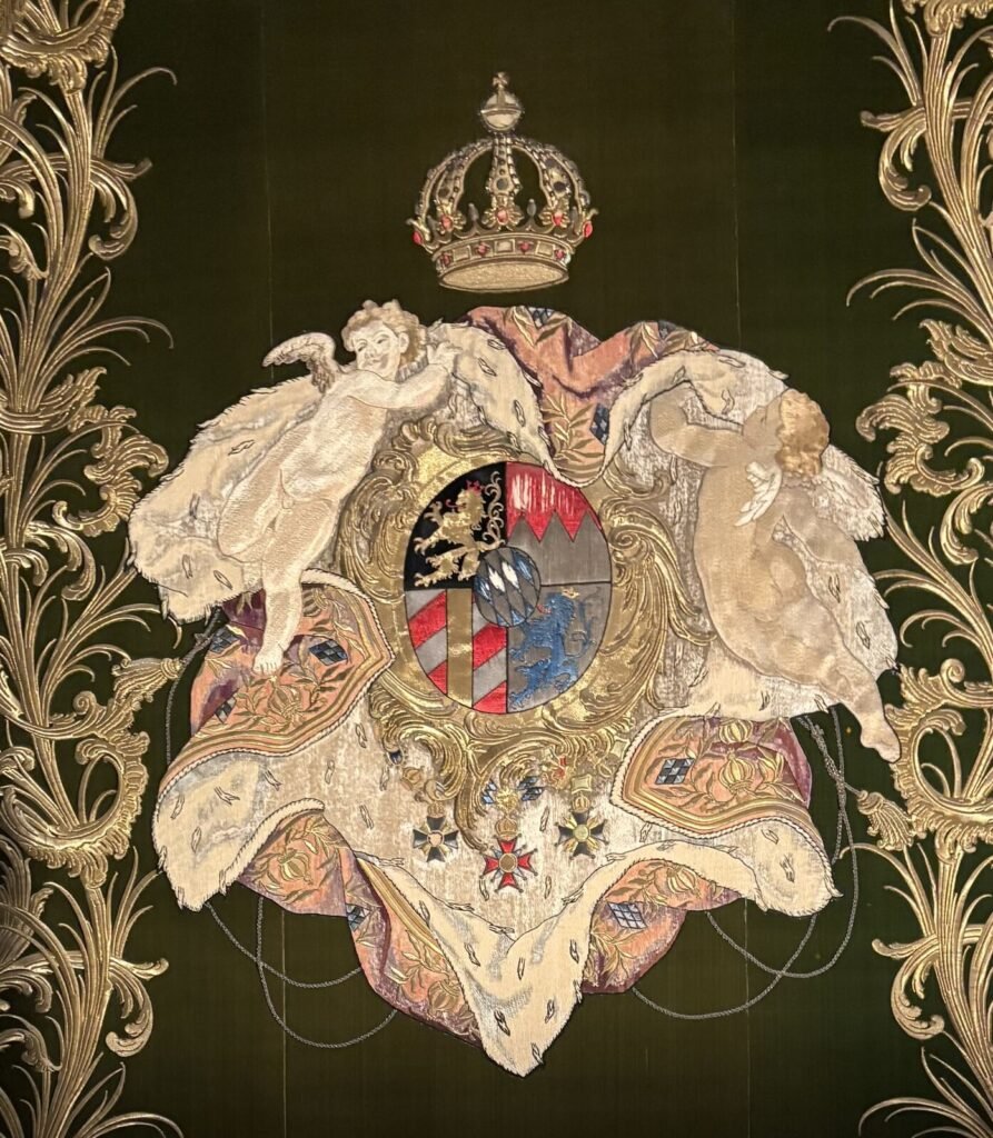 Ornate wall tapestry featuring a central coat of arms surrounded by decorative gold elements and cherubs, suggesting a Baroqu