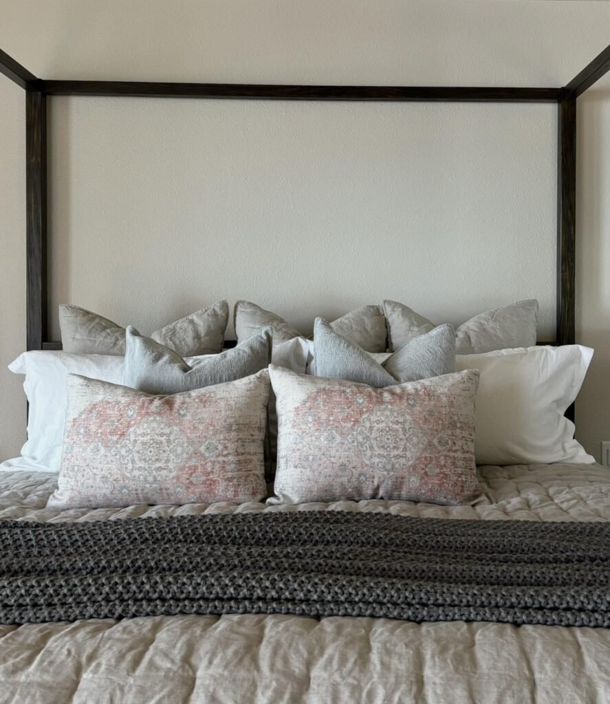 Cozy bedroom scene featuring a wooden four-poster bed with an array of decorative pillows in muted colors. The bedding has a
