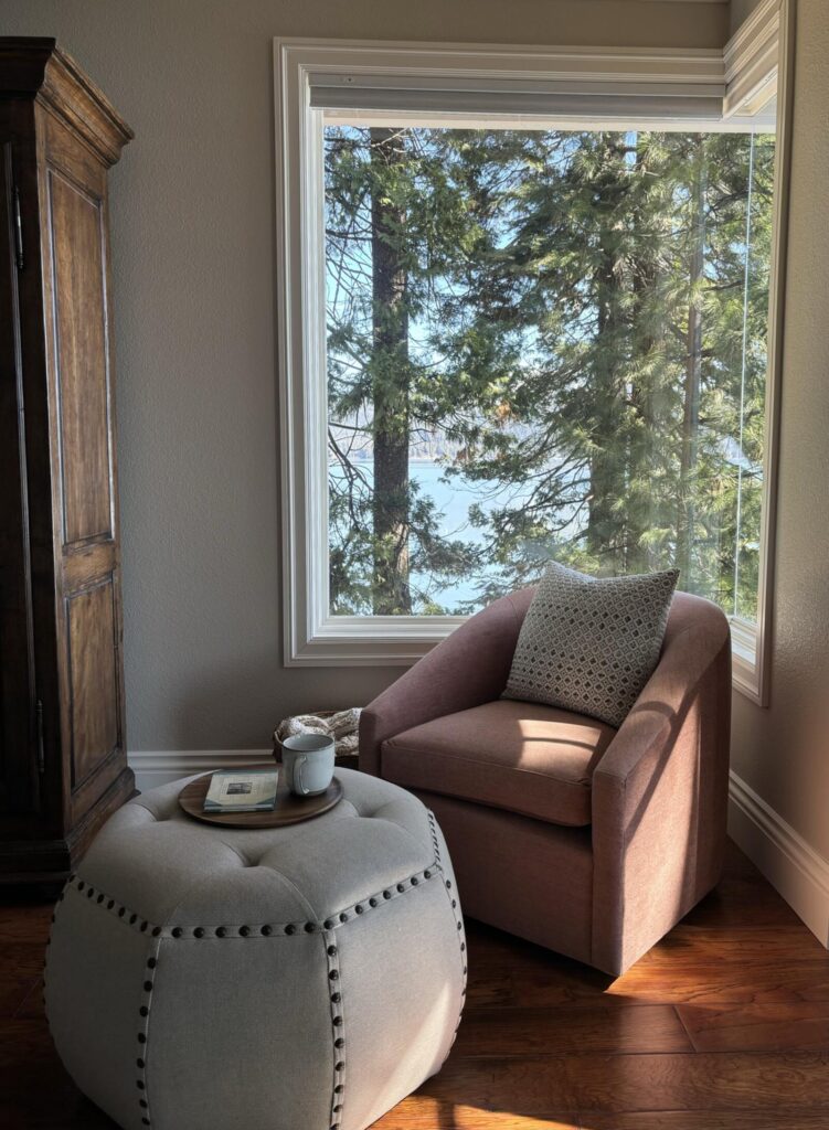 Cozy corner of a modern interior featuring a plush beige armchair and a round ottoman, with large windows overlooking a natur