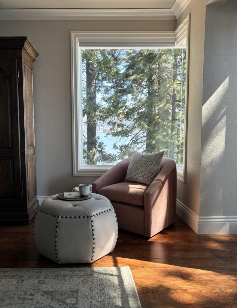 Cozy modern living room featuring a plush armchair and a round ottoman, with large windows revealing a serene lakeside view f