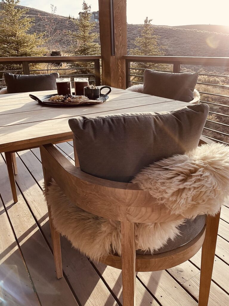 Modern rustic outdoor dining area featuring a wooden table and comfortable seating with plush, fur throw pillows. The scene i