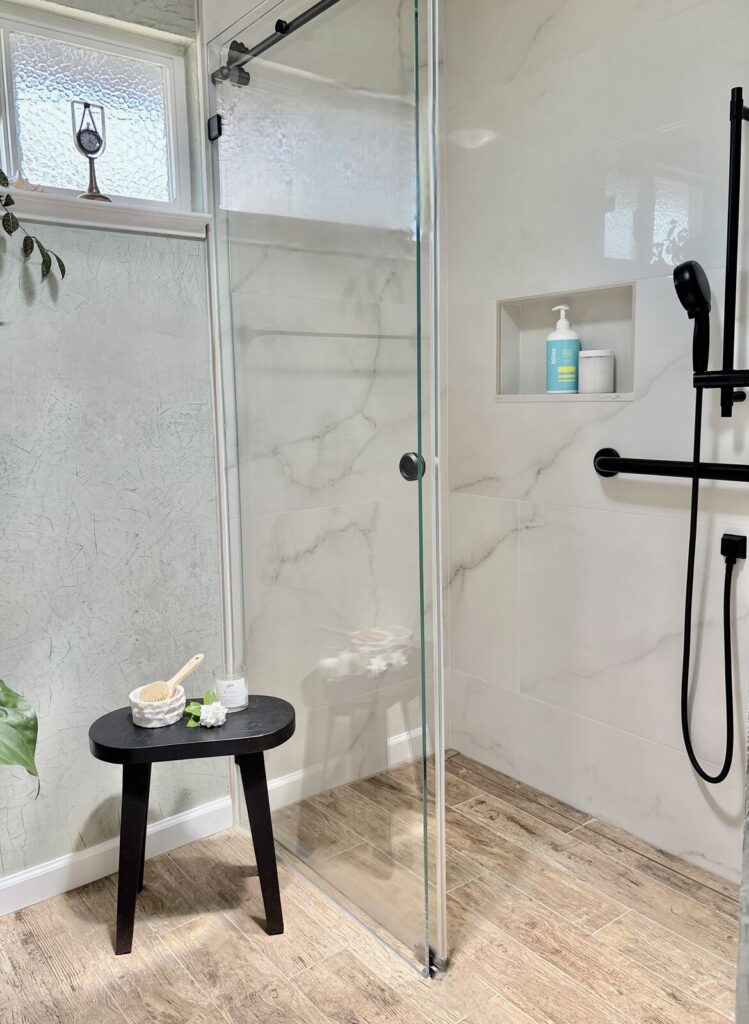 Modern bathroom with a glass shower enclosure, featuring marble walls and wood-look tile flooring. The space has a serene and
