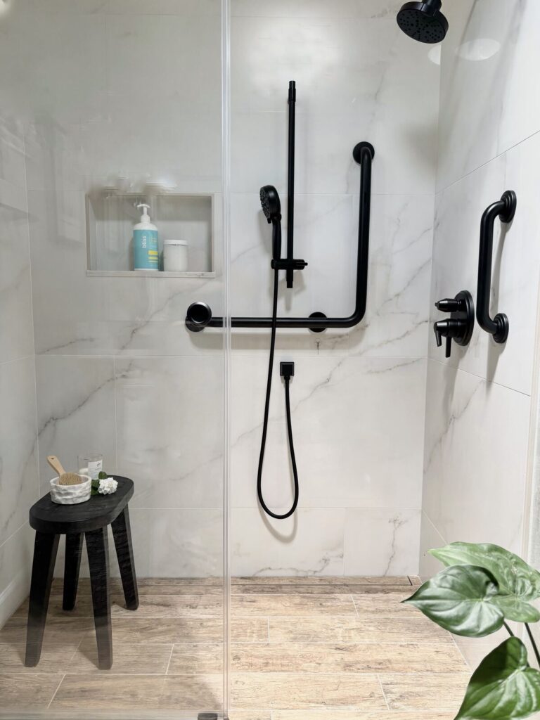 Modern bathroom shower with marble walls, black fixtures, and a small black stool. The design emphasizes accessibility featur