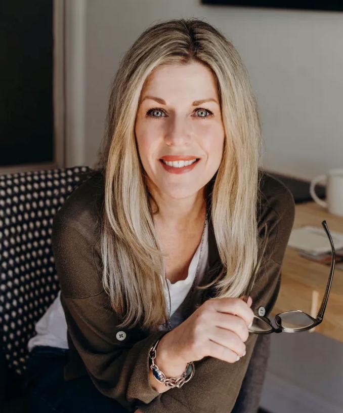 Woman with long blonde hair seated at a wooden table in a modern interior setting, wearing a casual outfit.