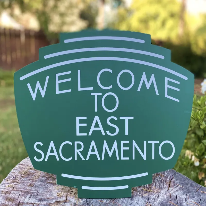 A green welcome sign for East Sacramento placed outdoors on a tree stump. — $50K Fabric And Upholstery Sacramento - Interior Designer