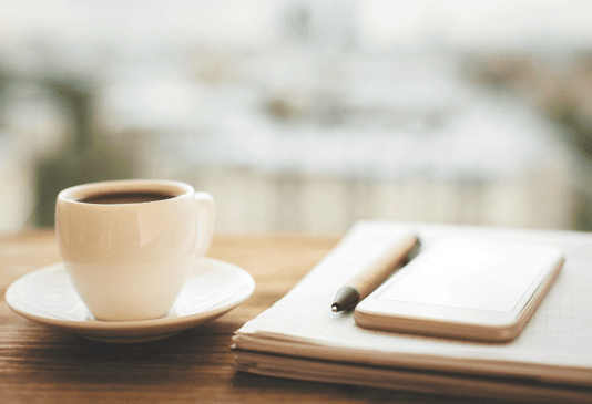 Contemporary workspace featuring a wooden table with a coffee cup, notebook, and smartphone, with a blurred cityscape in the