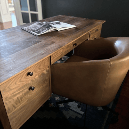 Modern home office setup featuring a wooden desk with multiple drawers and a sculptural, tan leather chair. The room has a da