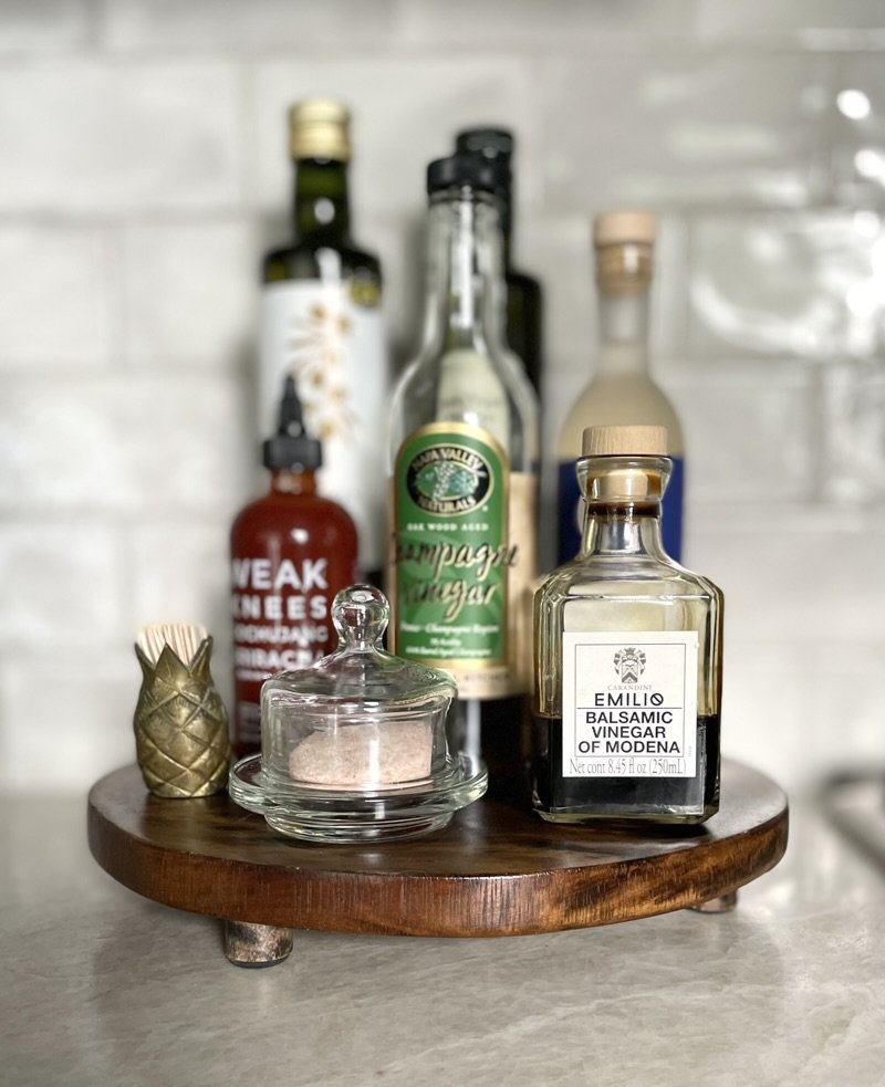 Rustic kitchen countertop featuring a wooden round board displaying various bottles of vinegar and sauces, with a glass conta