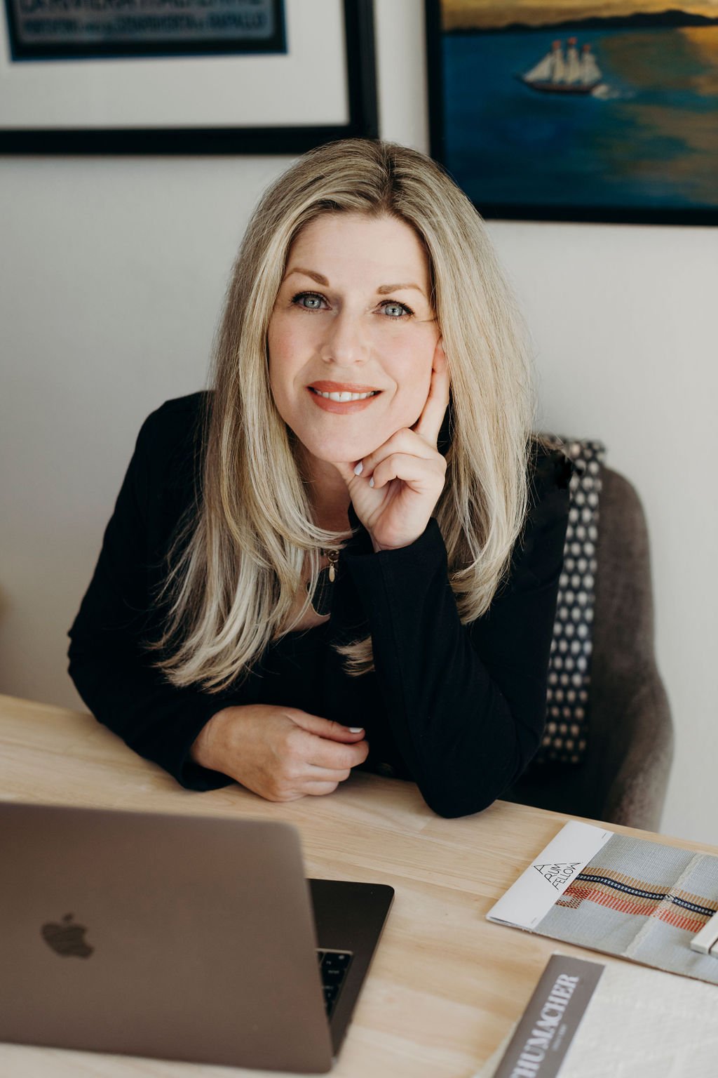 A woman with blonde hair sits at a wooden desk with a laptop and fabric samples, smiling and resting her chin on her