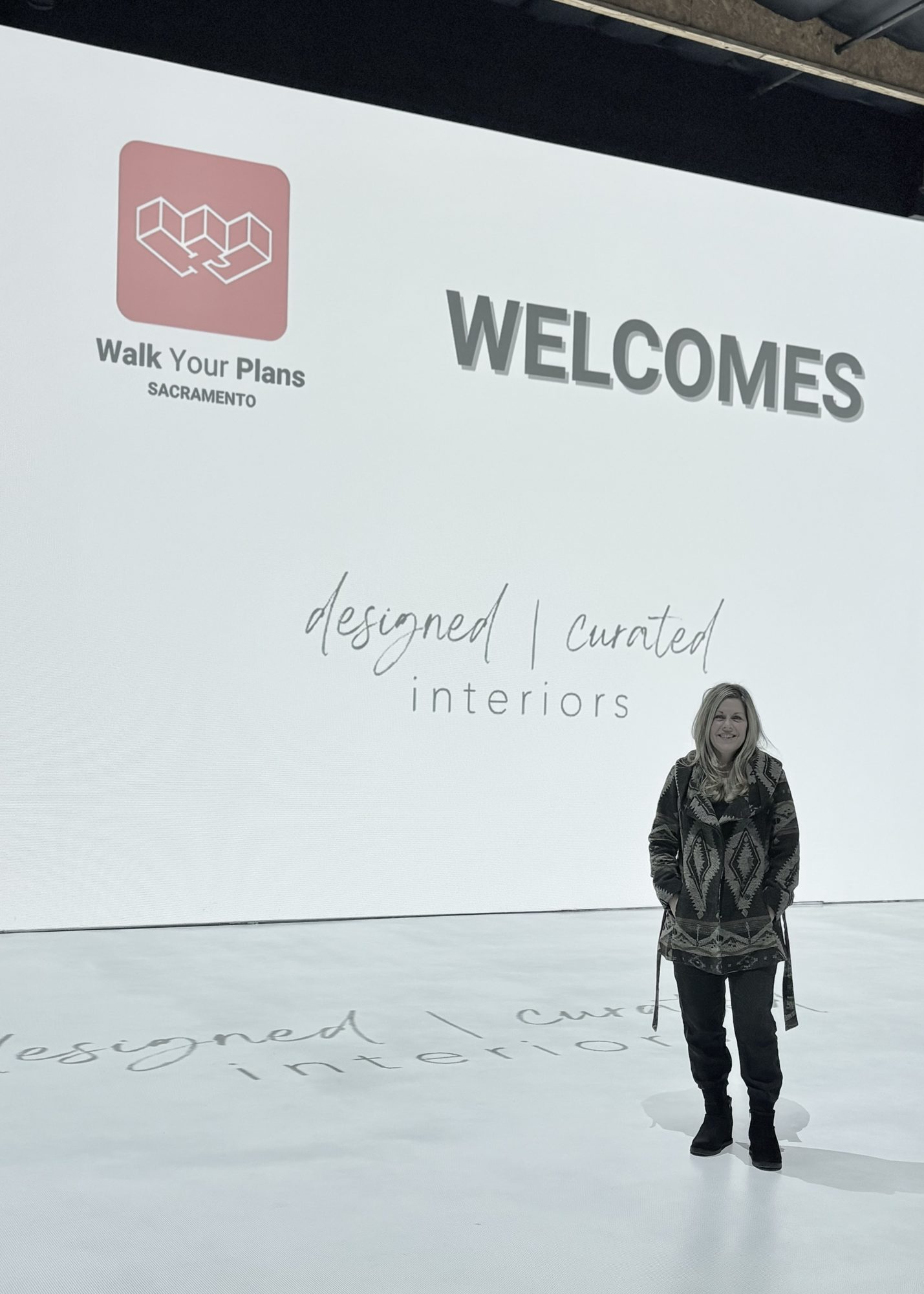 A woman stands in front of a large white wall with a logo and text welcoming to Walk Your Plans Sacramento, promoting de — Should I Hire Whole Home Renovation In Sacramento - Interior Designer
