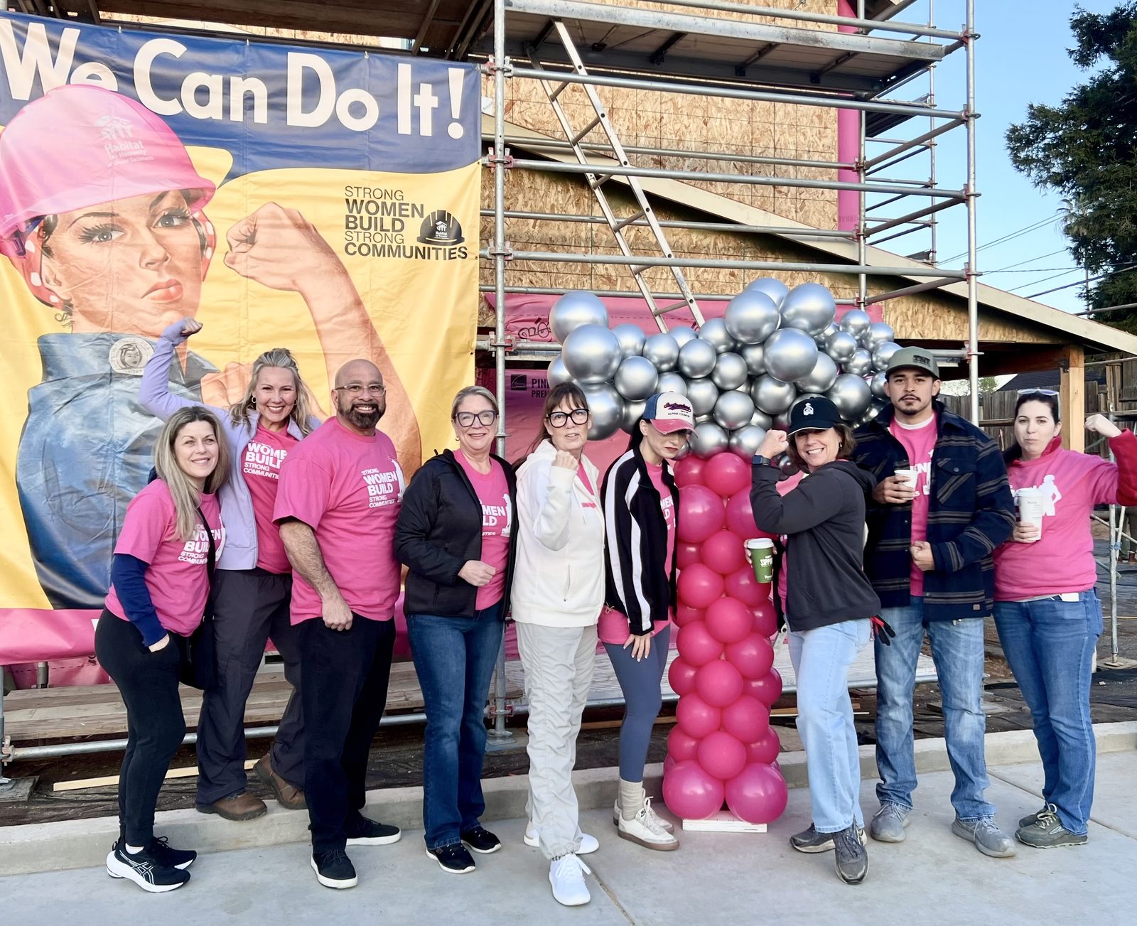 A group of people wearing pink shirts posing in front of a construction site with scaffolding and a large banner featuri — Should I Hire Whole Home Renovation In Sacramento - Interior Designer