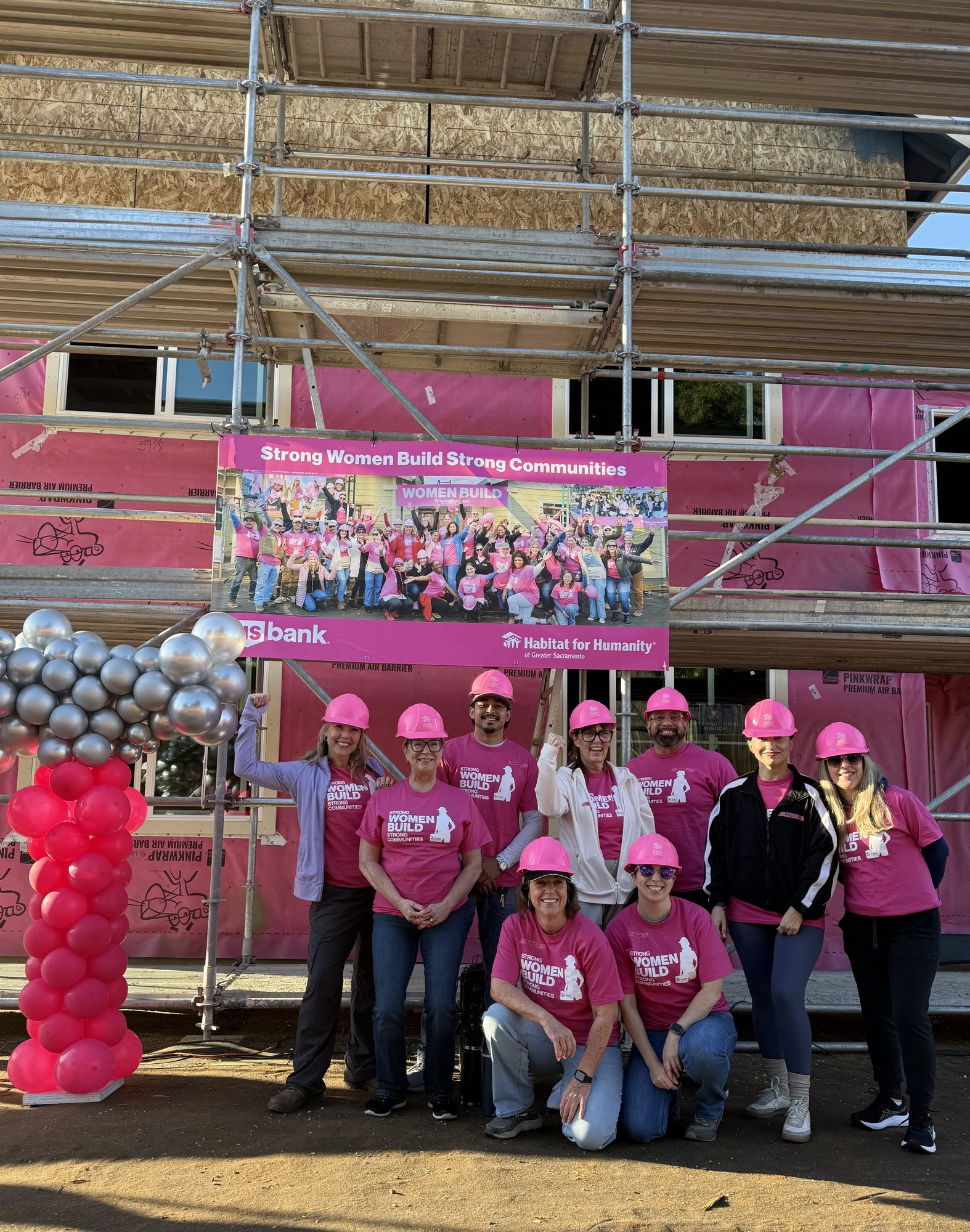 A group of people wearing pink hard hats and shirts posing in front of a construction site with scaffolding and a banner — How Much Does Structural Considerations For Master Suite Additions In Sacramento - Interior Designer