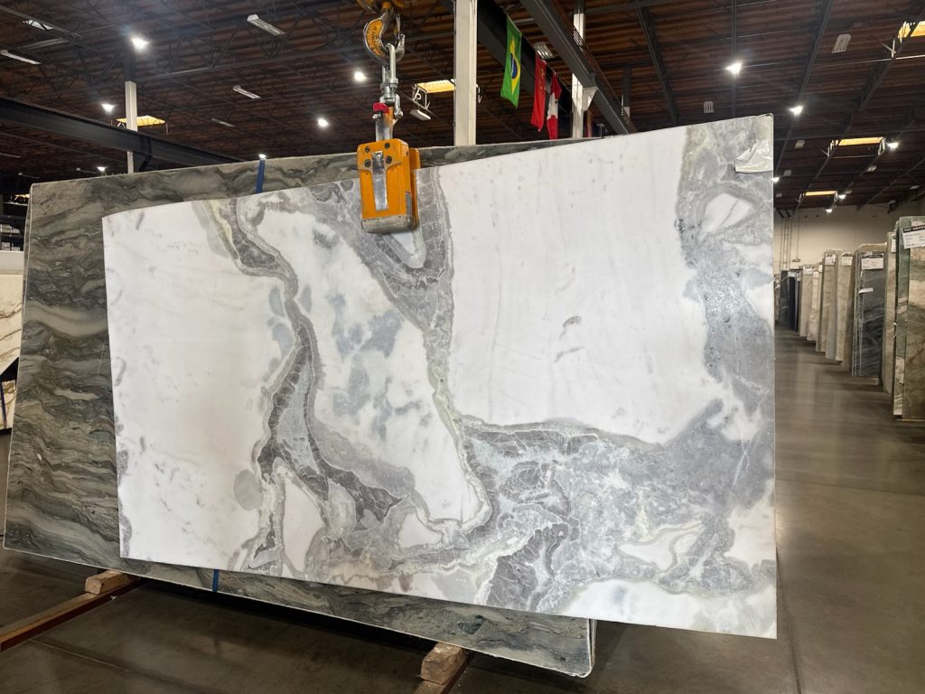Large slab of marble with a swirling gray and white pattern, showcasing a polished finish. The setting is a warehouse environ