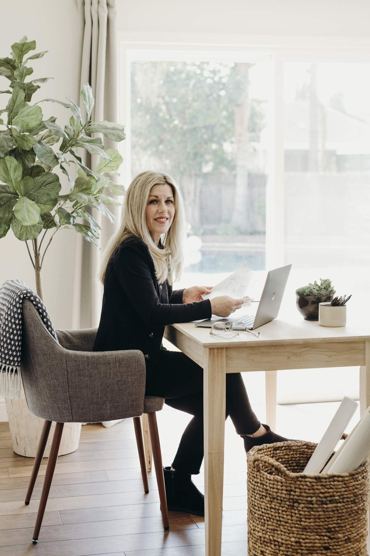 A woman sitting at a wooden desk in a bright, modern home office with a large window, a potted plant, and a woven basket