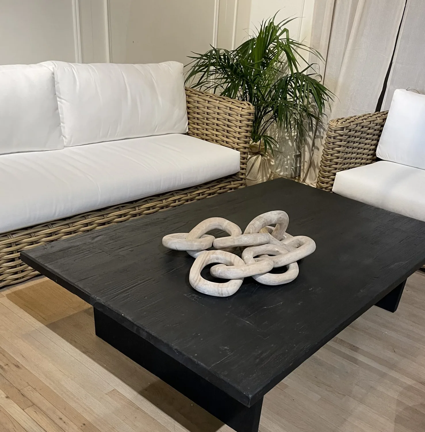 A living room with white cushioned wicker sofas, a black wooden coffee table, and a decorative chain sculpture