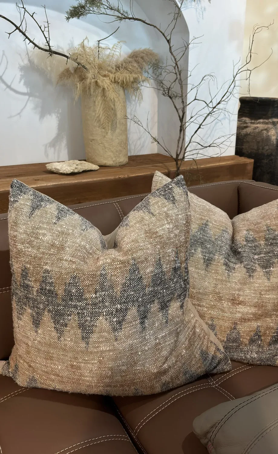 A close-up of a brown leather sofa with two textured patterned pillows, a wooden shelf behind it holding a large vase with dried branches and a stone