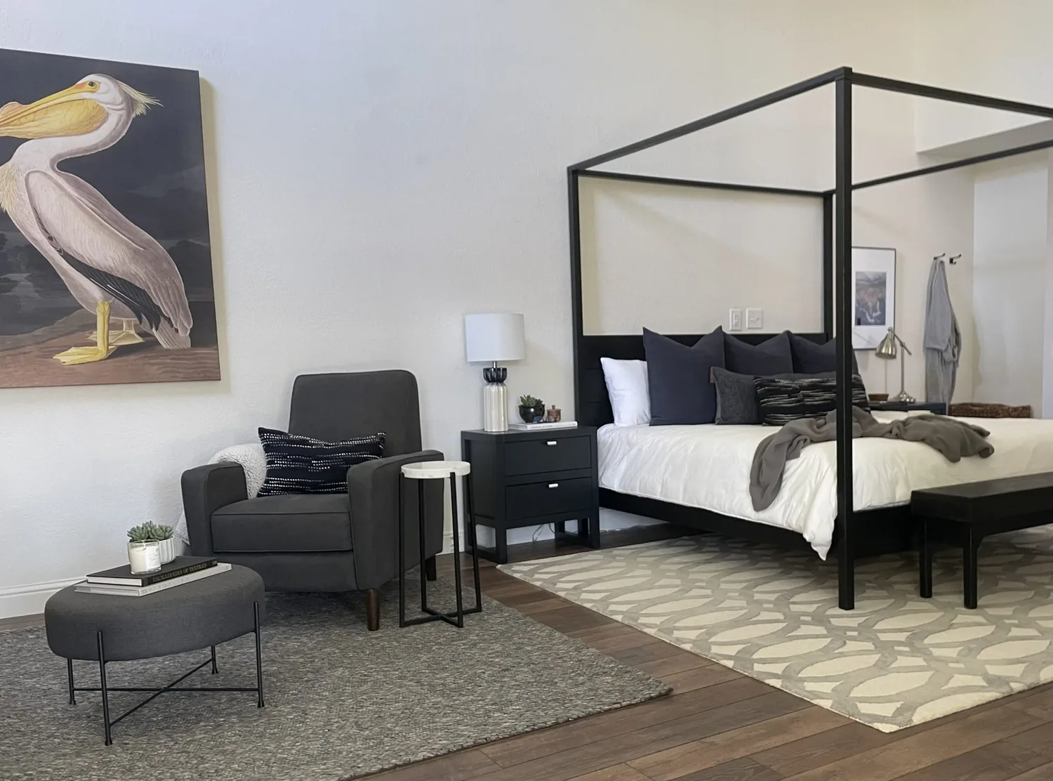 A neatly made bed with neutral bedding in a bright bedroom featuring a patterned area rug, framed photos above