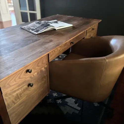 A wooden desk with drawers and a brown leather swivel chair in a room with a dark wall and a patterned rug