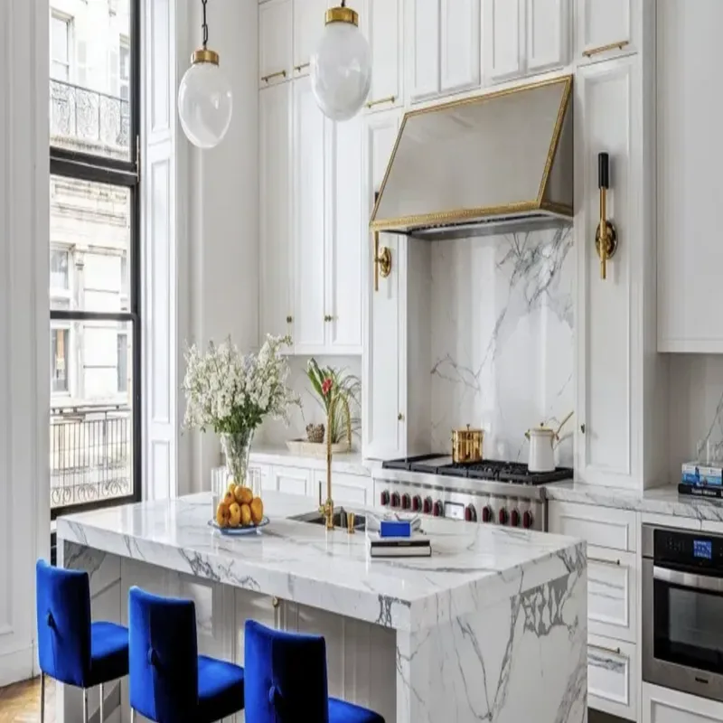 A modern kitchen with white marble countertops, white cabinetry, blue velvet chairs, and gold accents