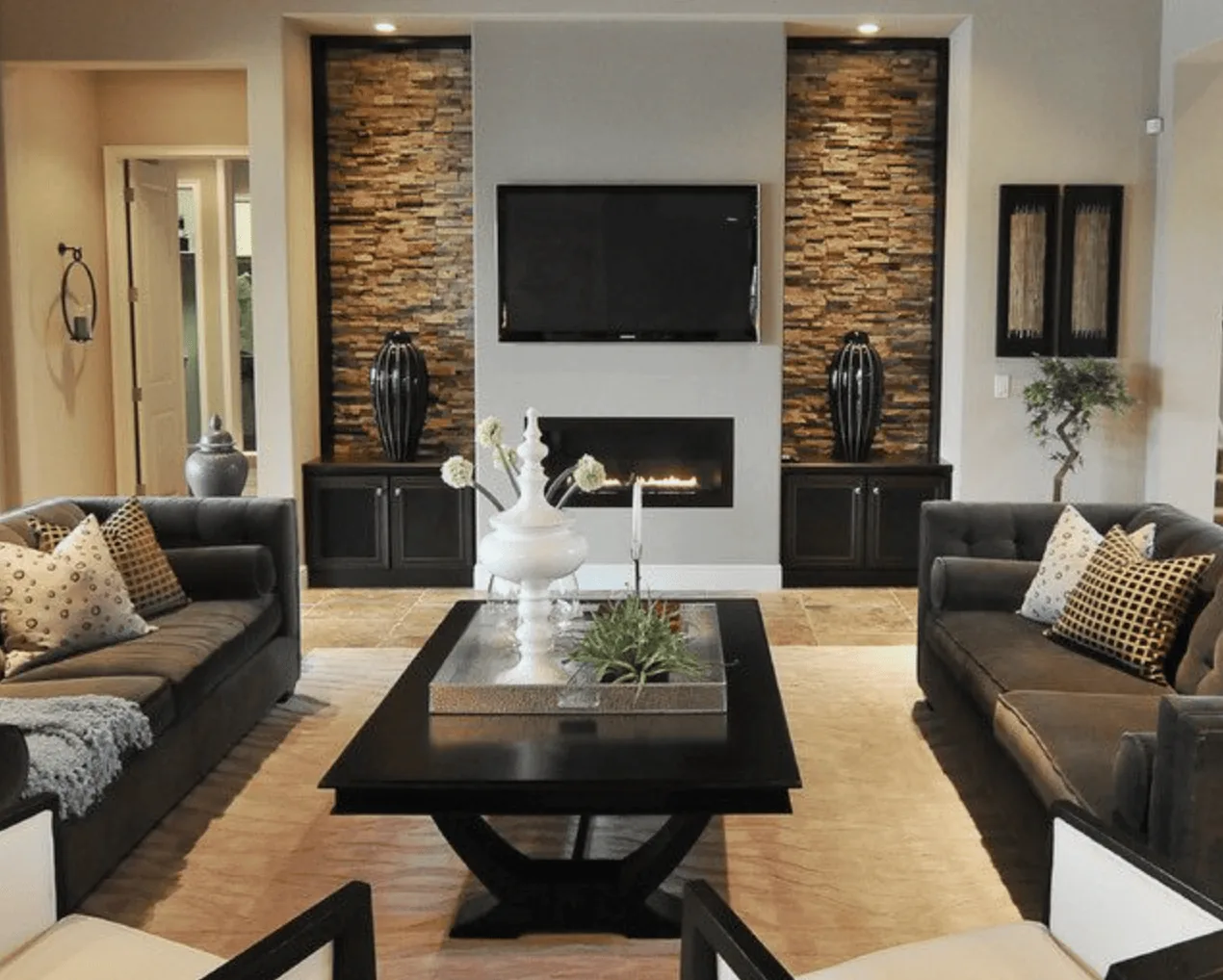 A modern living room with two dark sofas facing a black coffee table, a wall-mounted TV above a fireplace, and stone