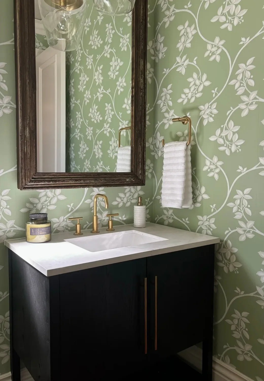 A bathroom vanity with a white countertop, gold fixtures, a dark wood cabinet, a large framed mirror, and green floral wallpaper