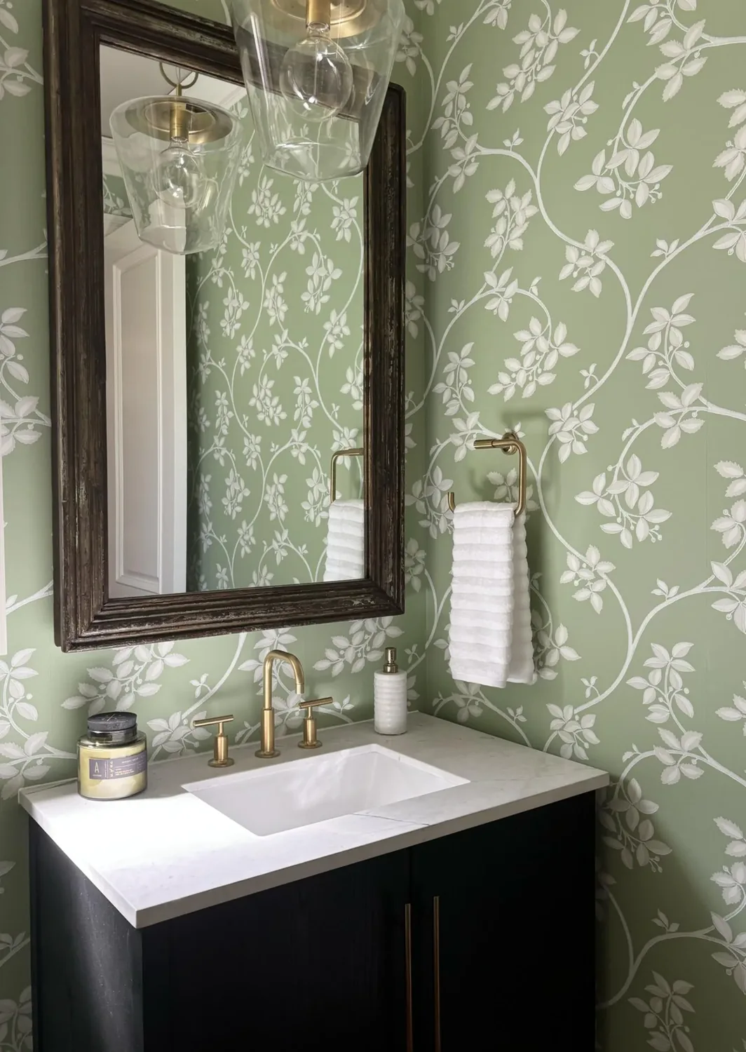 A bathroom vanity with a white sink, brass fixtures, a dark wood cabinet, a large framed mirror, and green floral