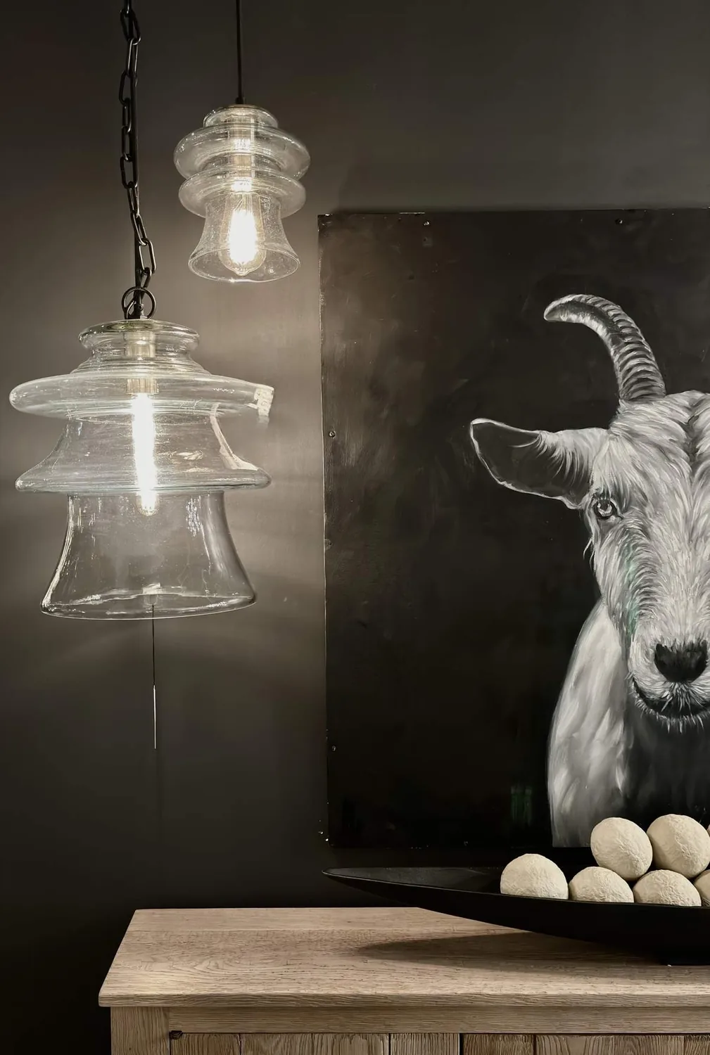 A modern interior scene featuring two clear glass pendant lights, a wooden table, a black bowl with decorative white spheres, and a black and white painting of a goat on a dark wall