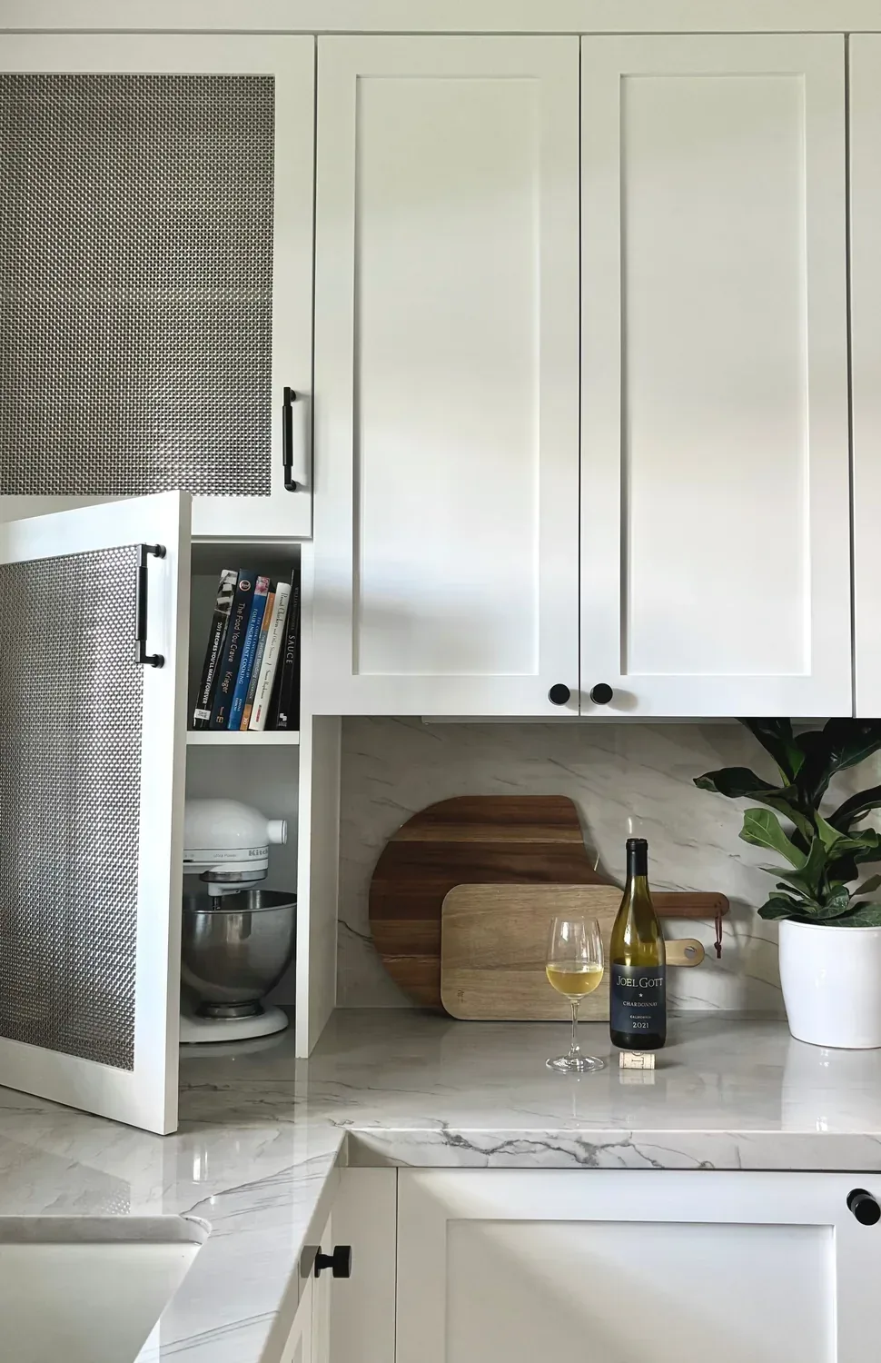 A modern kitchen countertop with white cabinets, a wine bottle and glass, cutting boards, a plant, and a mixer inside