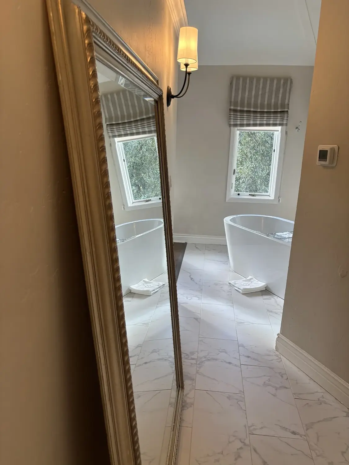 A bathroom with a freestanding bathtub, marble tile floor, a window with striped curtains, and a wall-mounted lamp reflected in a large mirror