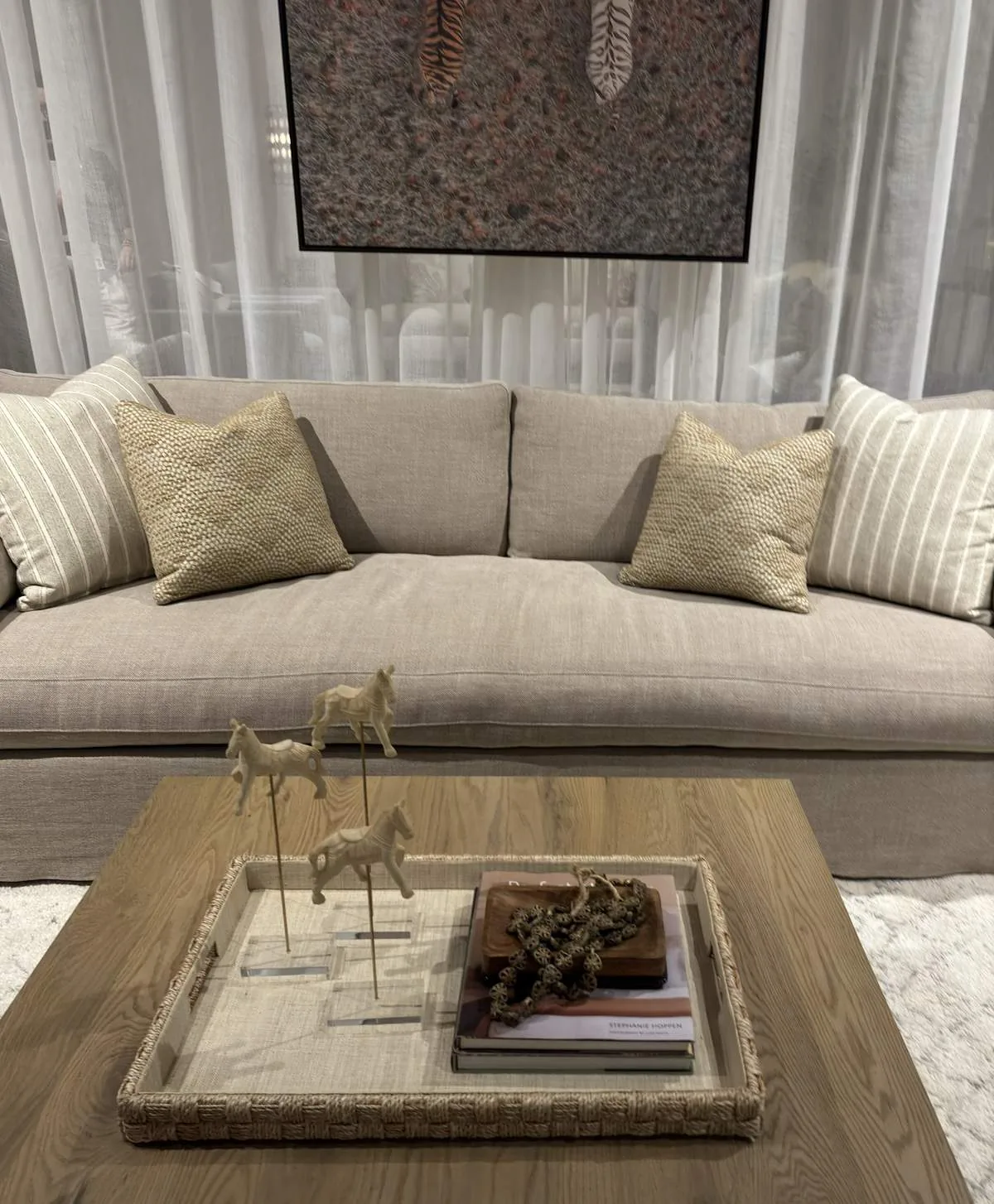 A beige sofa with textured and striped pillows in front of sheer curtains, with a wooden coffee table holding a woven