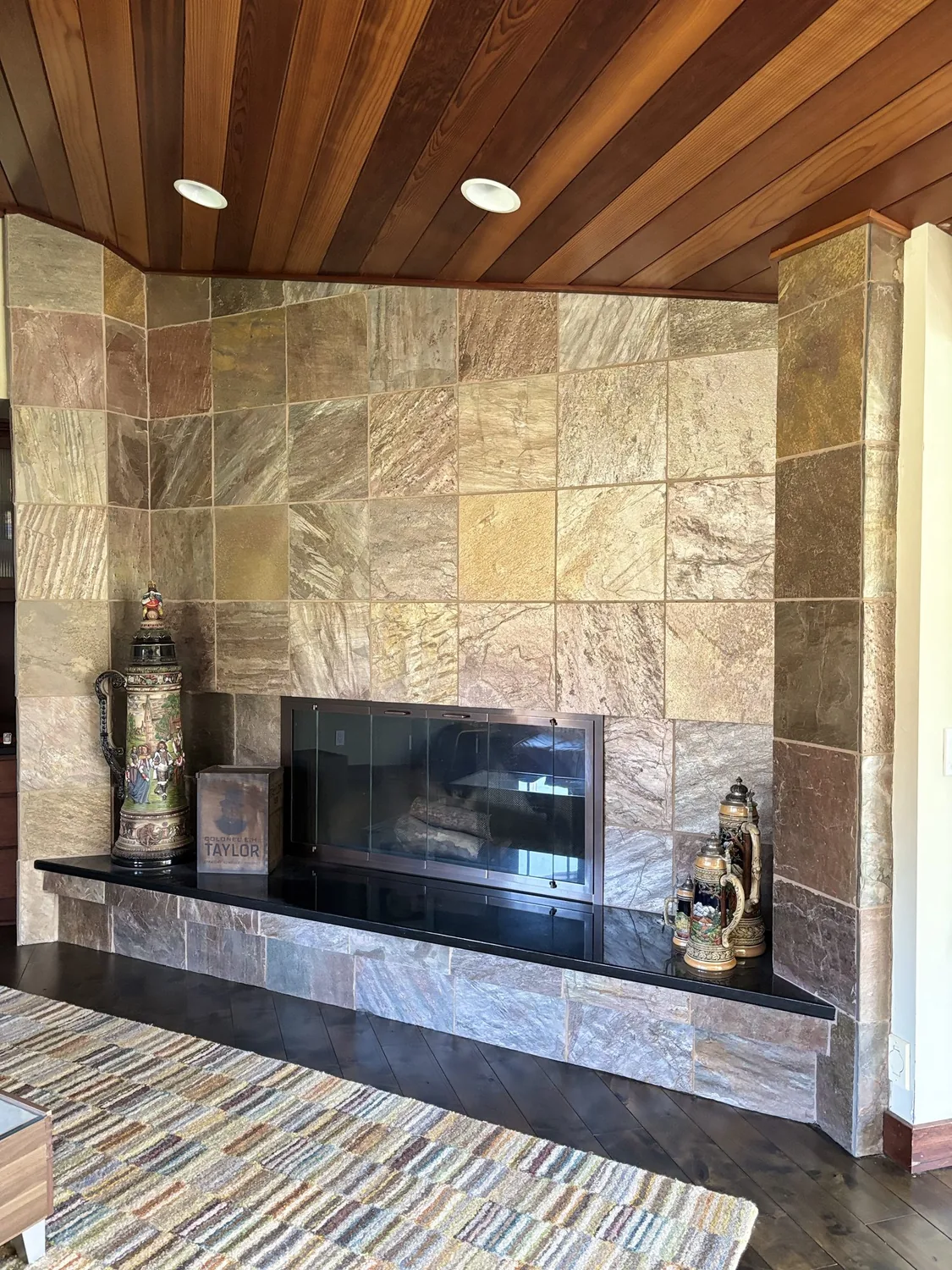 A living room fireplace with a stone tile surround, a black hearth, wooden ceiling, and decorative steins on the hearth