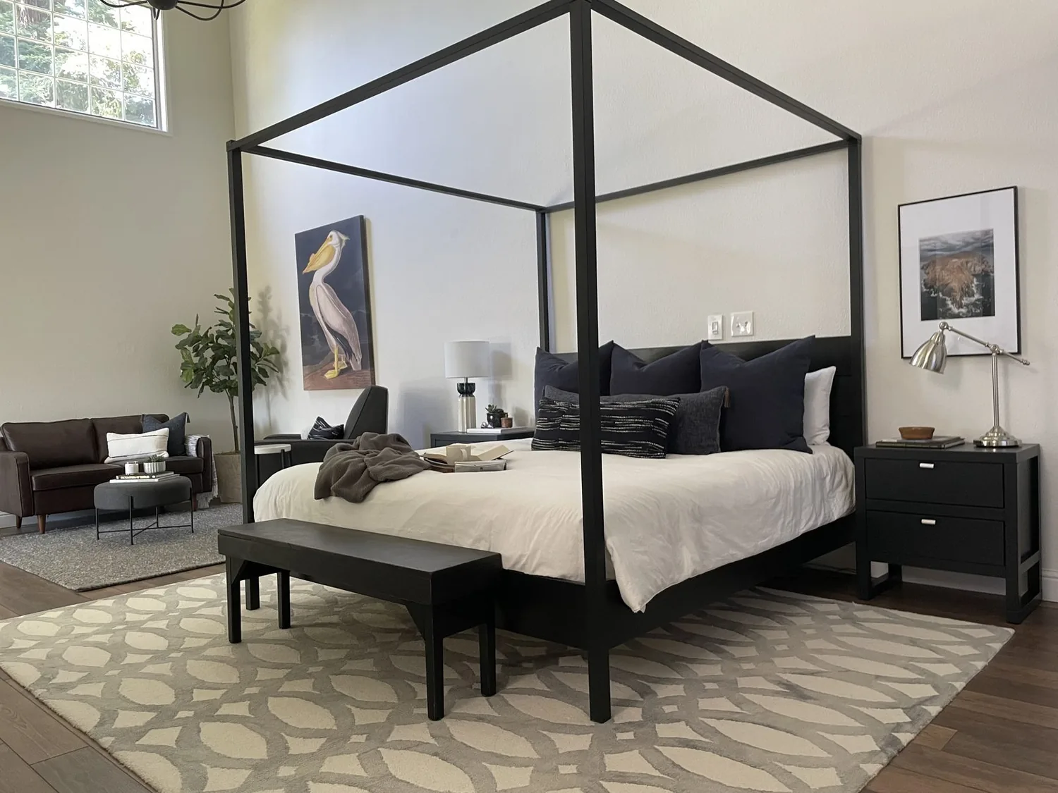 A modern bedroom with a black four-poster bed, neutral bedding, a patterned rug, and a seating area with a brown