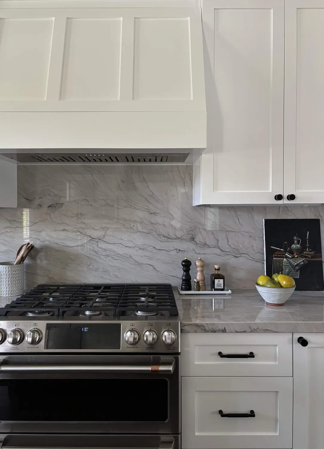 A modern kitchen featuring a stainless steel gas stove, white cabinetry, marble backsplash and countertop, with a bowl of pears and kitchen utensils on the counter