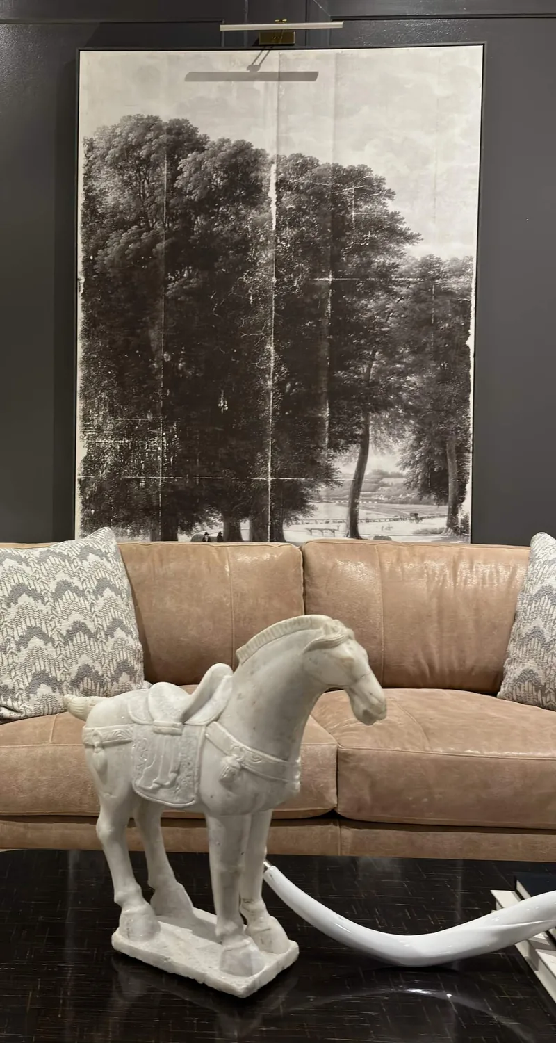 A living room with a tan leather sofa, patterned pillows, a large black and white tree artwork on the wall, and a white decorative horse statue on a black table