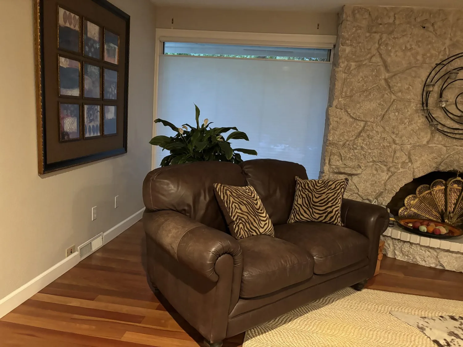 A living room with a brown leather loveseat, zebra print pillows, a stone fireplace, a large plant, and a framed