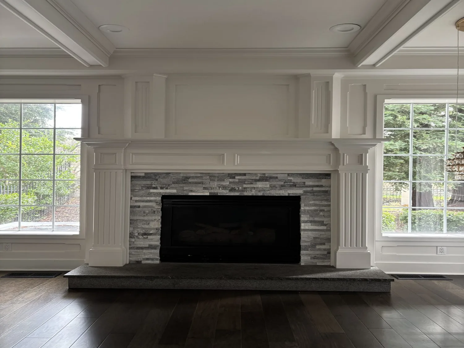 A living room fireplace with white detailed molding, gray stone surround, and large windows on either side
