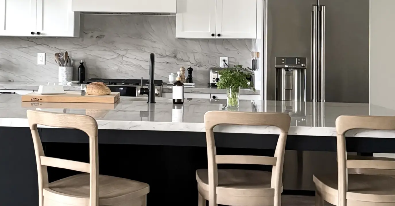 A modern kitchen with a marble countertop island, light wood chairs, white cabinets, and stainless steel appliances