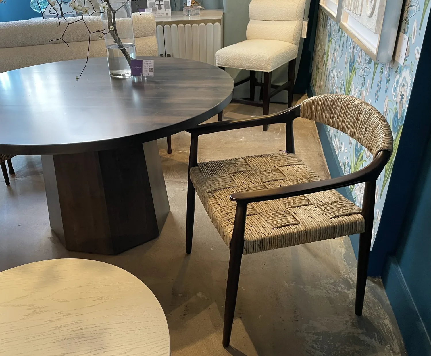 A dining area with a round wooden table, a woven seat chair, a cushioned chair, and floral wallpaper on the wall