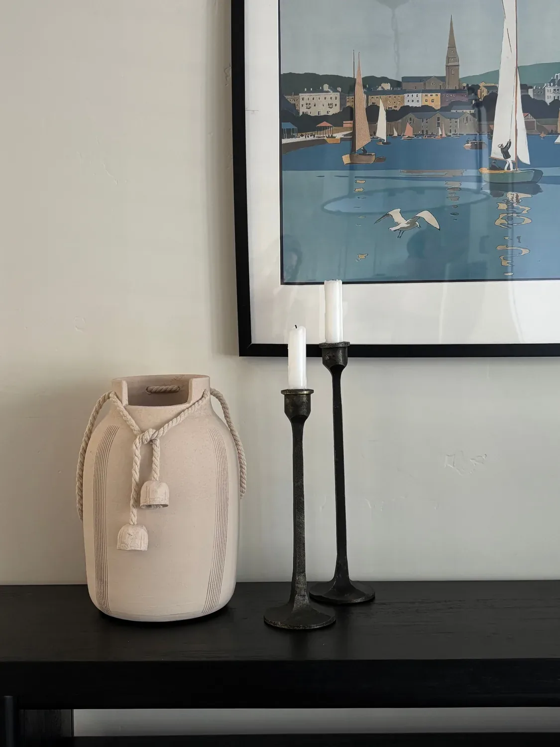 A minimalist interior vignette featuring a beige ceramic vase with rope detail, two black metal candlesticks with white