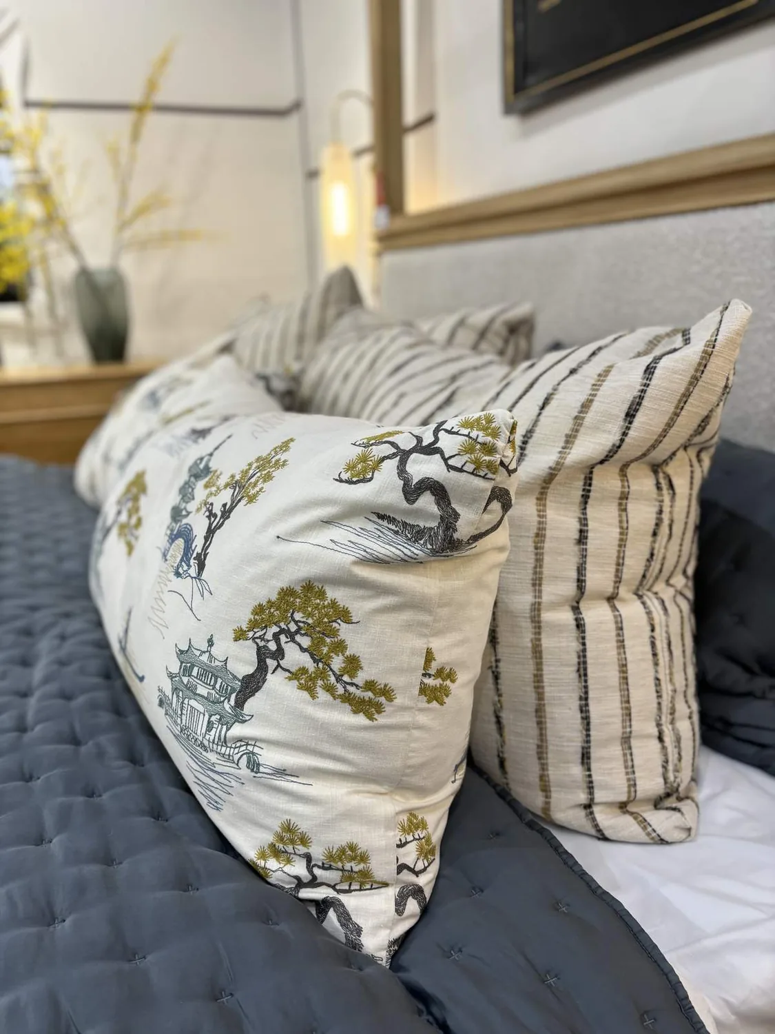 Close-up of decorative pillows on a bed with a tufted dark gray comforter and a blurred background featuring a vase with yellow flowers
