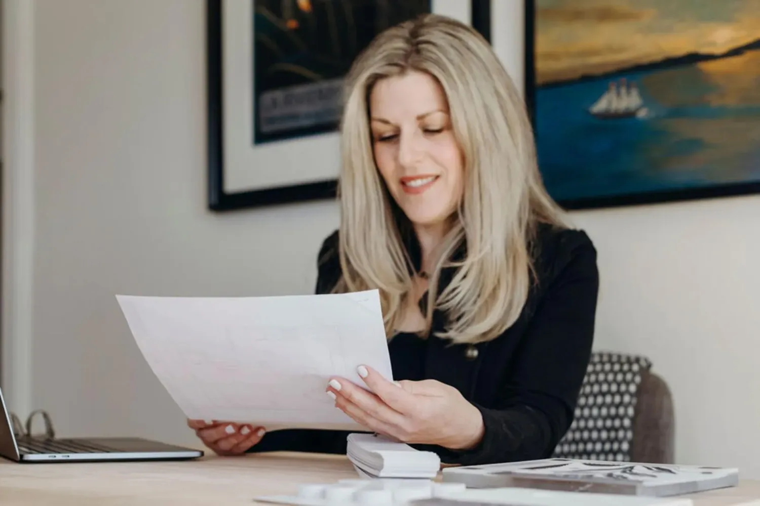 A woman sitting at a desk reviewing papers with a laptop and books nearby, in a room with framed artwork on the wall