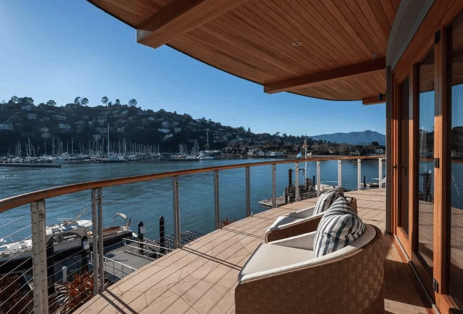 A curved balcony with wicker chairs and striped cushions overlooking a marina with boats and hills