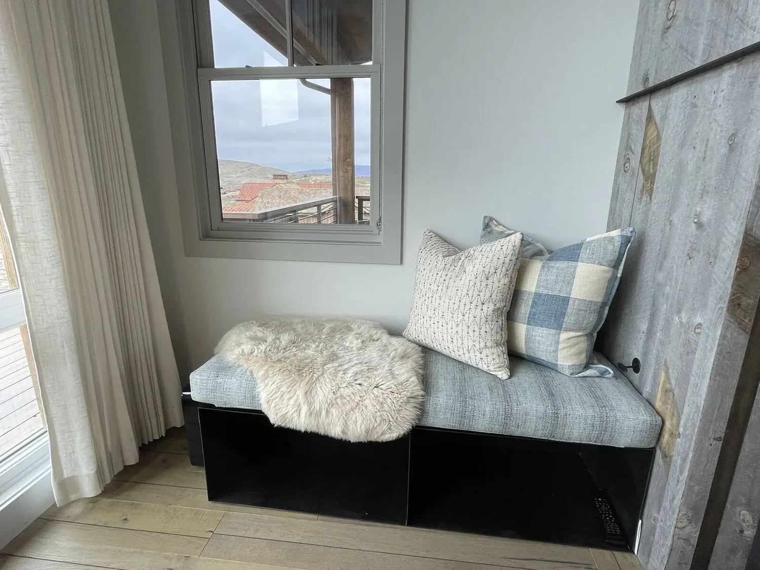 A cozy window nook with a cushioned bench, two pillows, and a faux fur throw, next to a window with a view of rooftops